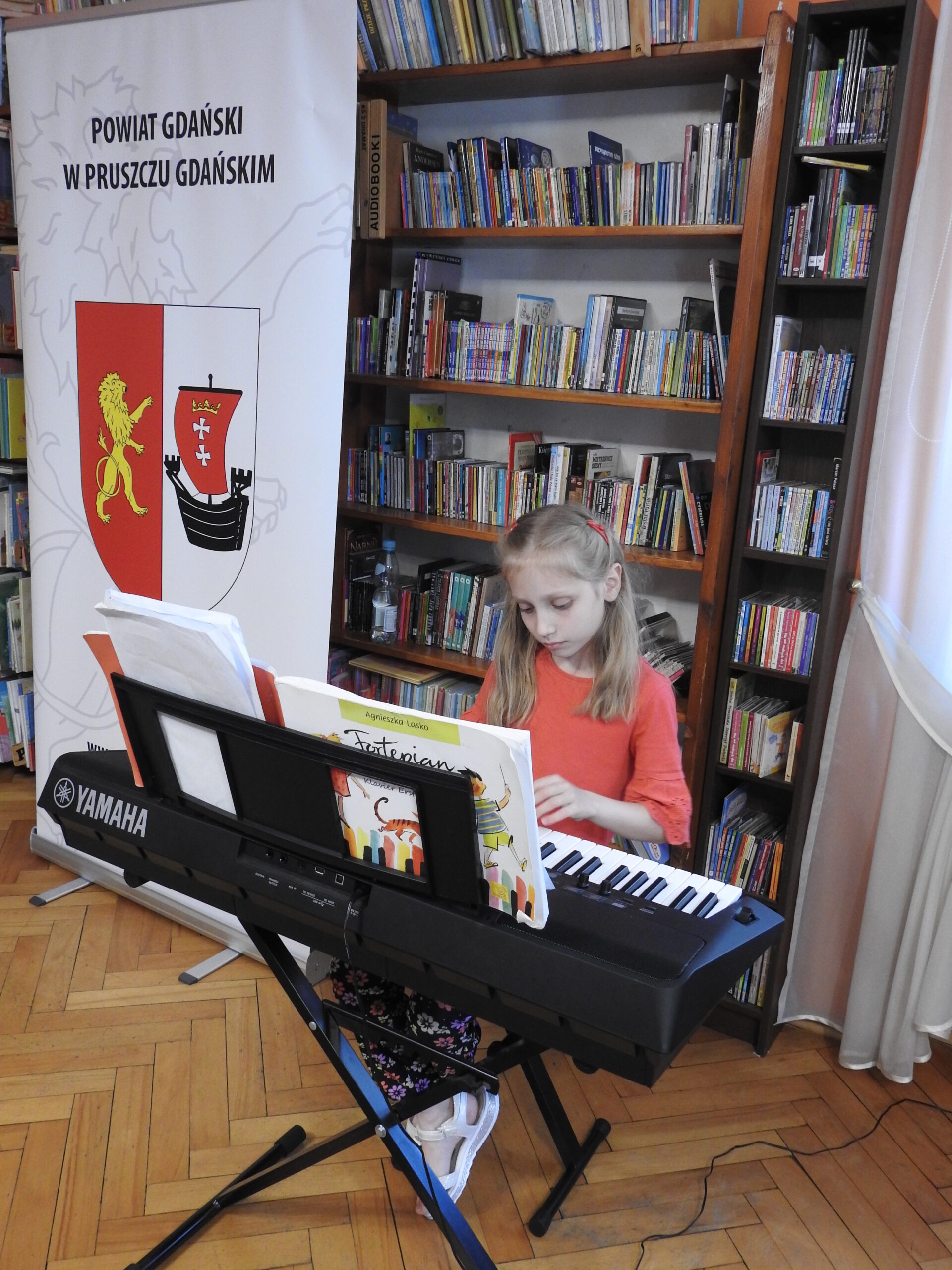 Dziewczynka siedzi przy instrumencie klawiszowym firmy Yamaha i trzyma dłonie nad klawiszami. Na instrumencie znajdują się otwarte nuty. W tle regały z książkami oraz baner z napisem Powiat Gdański w Pruszczu Gdańskim oraz herbem Powiatu Gdańskiego.