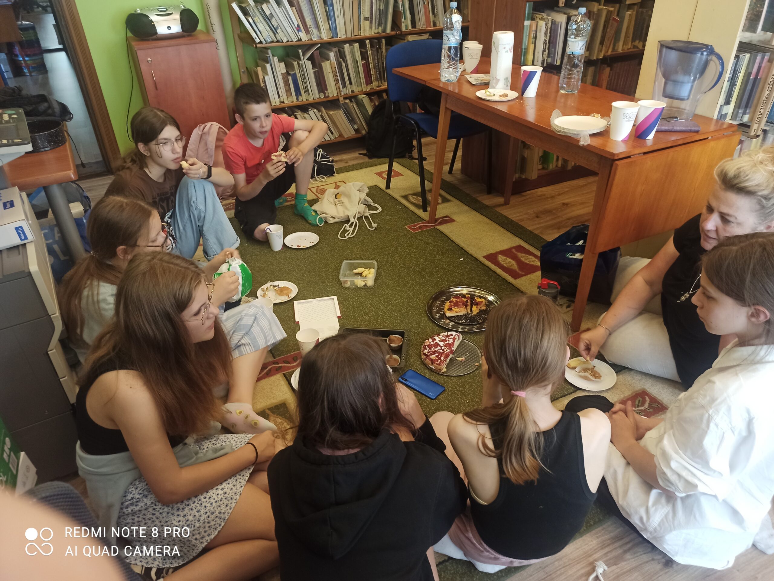 Dzieci z Dyskusyjnego Klubu Książki wraz z moderatorką siedzą na dywanie w okręgu. Na dywanie postawione są papierowe talerzyki, kubeczki, ciasta, rogaliki, babeczki.