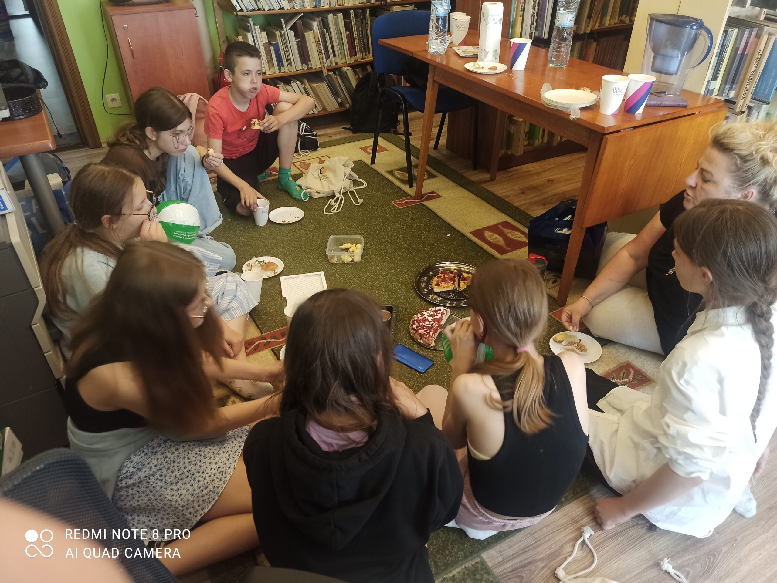 Grupa dzieci z Dysksyjnego Klubu Książki siedzi na dywanie i rozmawia o książce. Niekóre dzieic jedzą przyniesione słodycze.
