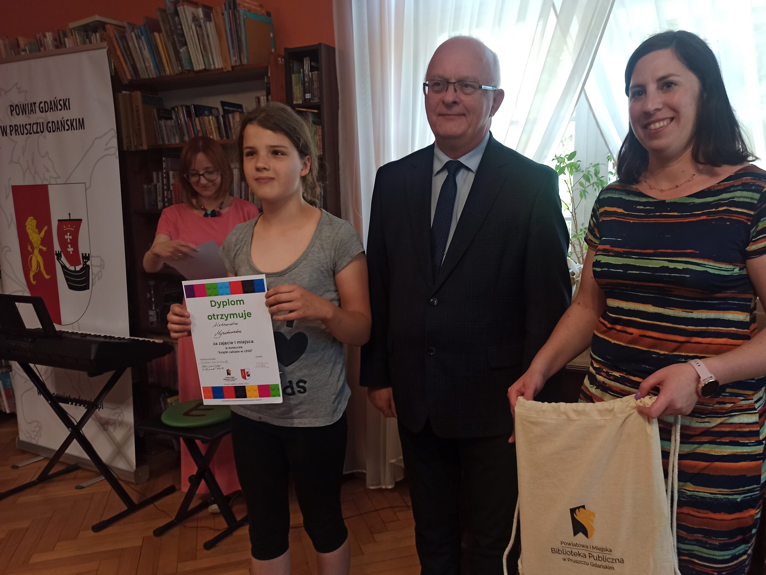 Dziewczynka oraz Starosta Marian Cichon i Natalia Błońska z Zarządu Powiatu Gdańskiego stoją razem i uśmiechają się. Dziewczynka trzyma w rękach dyplom oraz torbę materiałową. W tle regały z książkami.