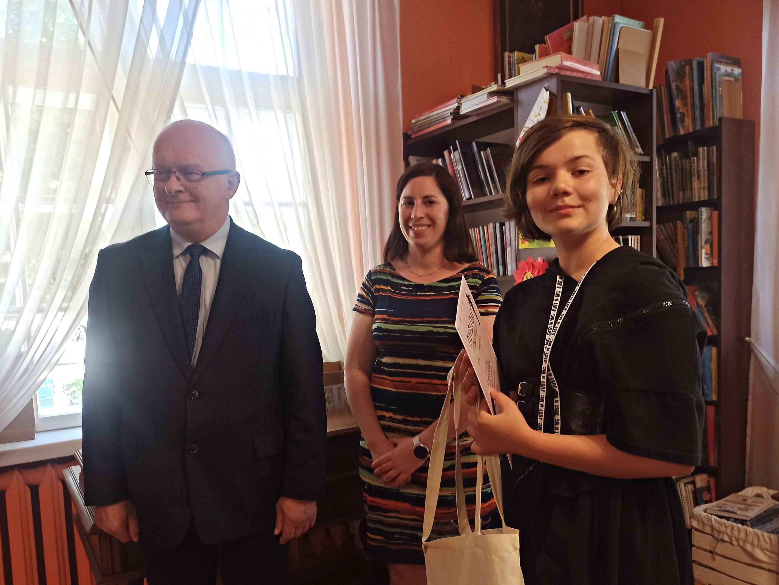 Dziewczynka oraz Starosta Marian Cichon i Natalia Błońska z Zarządu Powiatu Gdańskiego stoją razem i uśmiechają się. Dziewczynka trzyma w rękach dyplom oraz torbę materiałową. W tle regały z książkami.