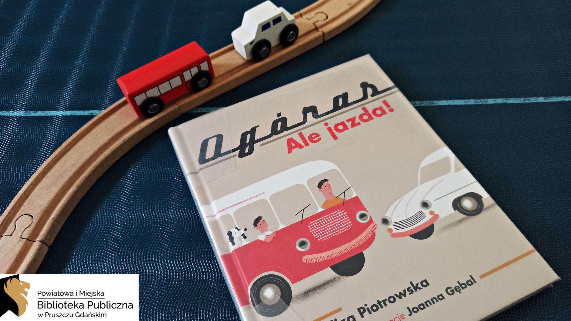 Na granatowym tle leży książka pt. „Ogóras. Ale jazda!”. Na okładce znajduje się zabytkowy czerwony autobus oraz biały samochód. Obok książki leży drewniany tor, na którym stoją dwa drewniane pojazdy – również czerwony autobus i biały samochód.