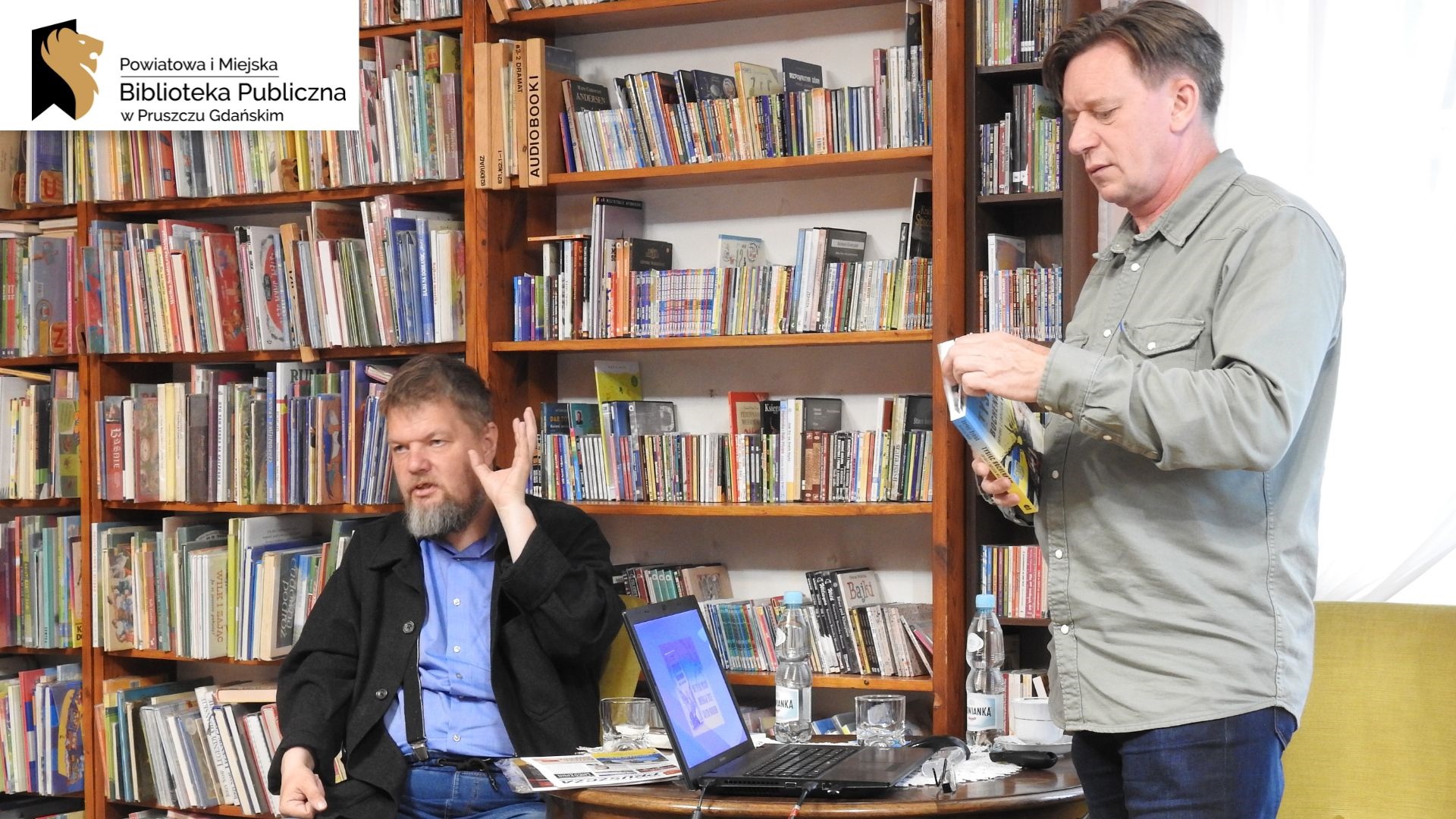 Serhyj Syniuk siedzi na fotelu z uniesioną dłonią, a Krzysztof Petek stoi trzymając książkę pt. Tysiąc bochenków. Pomiędzy nimi znajduje się stół, na którym jest włączony laptop, dwie butelki wody mineralnej, dwie szklanki oraz gazeta. W tle regały z książkami.