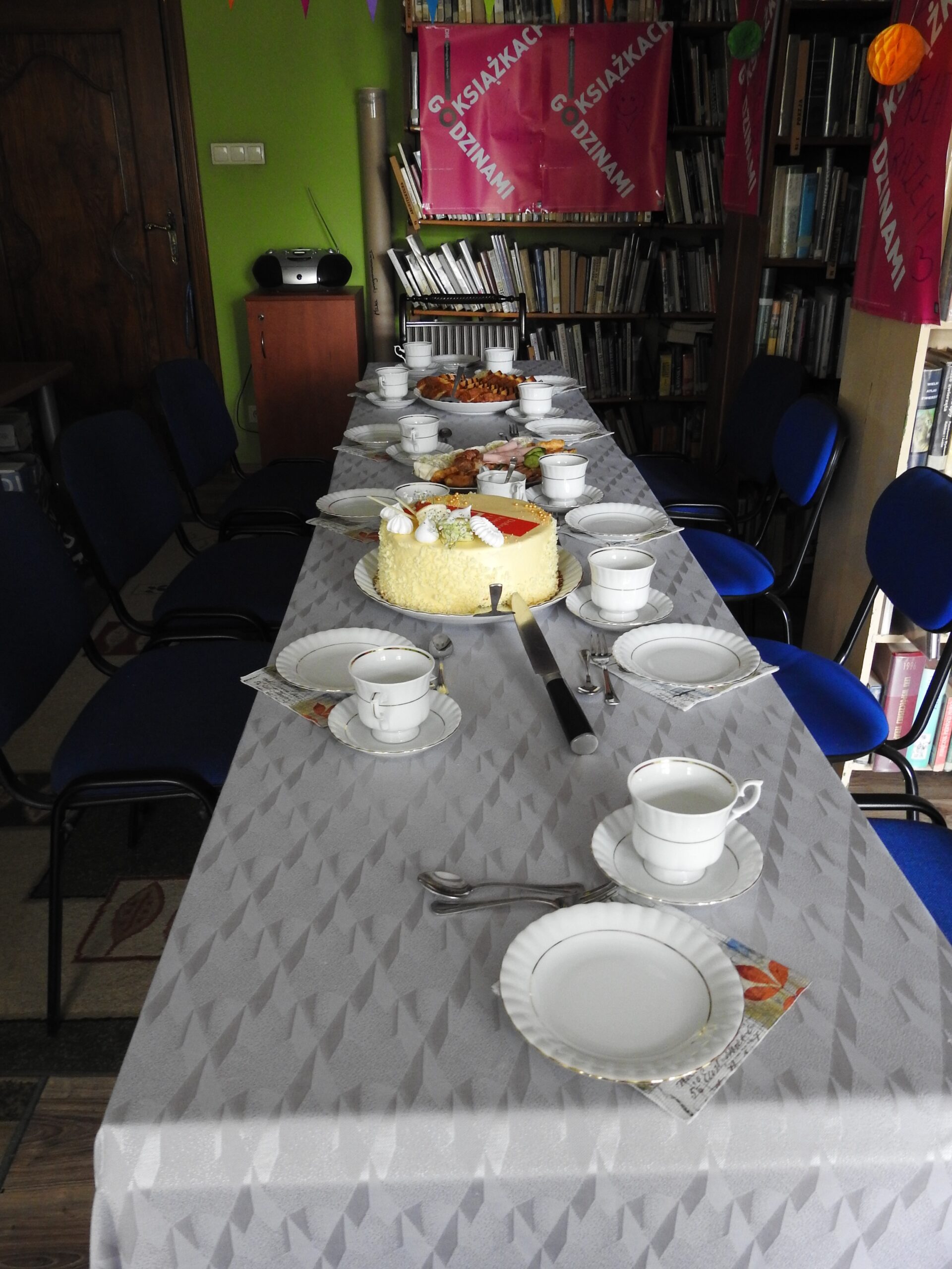 Stół nakryty srebrnym obrusem, na którym stoją filiżanki i talerzyki oraz tort, ciasta oraz półmisek z mięsnymi przekąskami. 