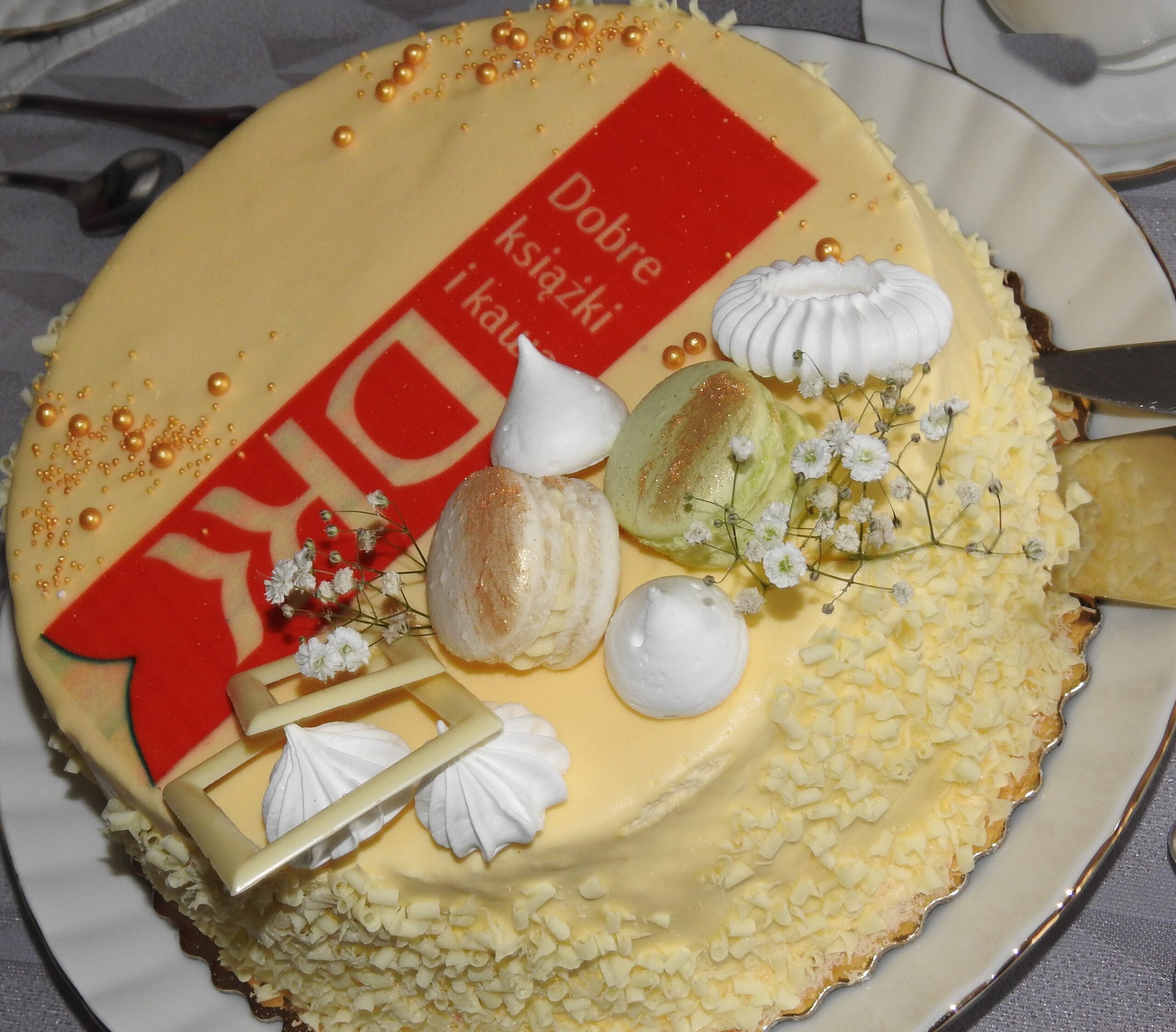 Tort ozdobiony bezami, białą czekoladą, złotymi kuleczkami oraz różową zakładką do ksiązki, na której widnieje napis: Dobre ksiązki i kawa, DKK.