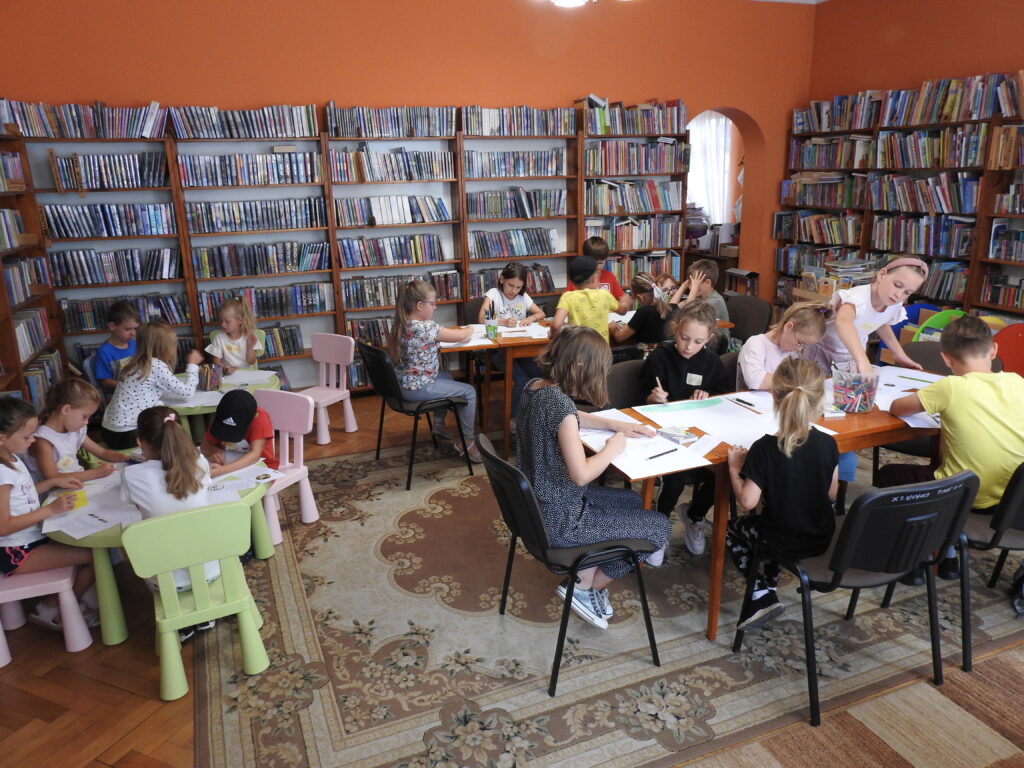 Duża grupa dzieci siedzi przy czterech stołach. Każde dziecko rysuje kredkami na białych kartkach formatu A3. W tle regały z książkami.