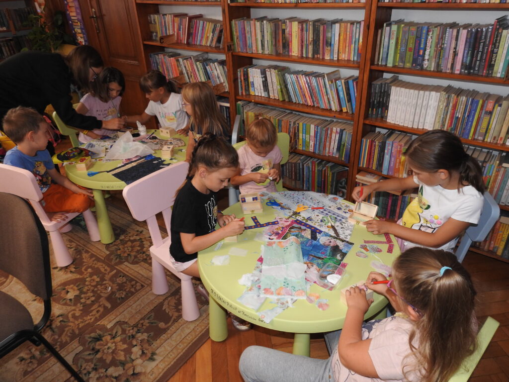 Wokół dwóch okrągłych stolików siedzi grupa dzieci. Przed nimi rozłożone są kolorowe papierowe serwetki, pędzelki, pudełeczka oraz opakowanie kleju. Dzieci wycinają oraz malują pędzelkami po  małych drewnianych pudełeczkach.  W tle regały z książkami.
