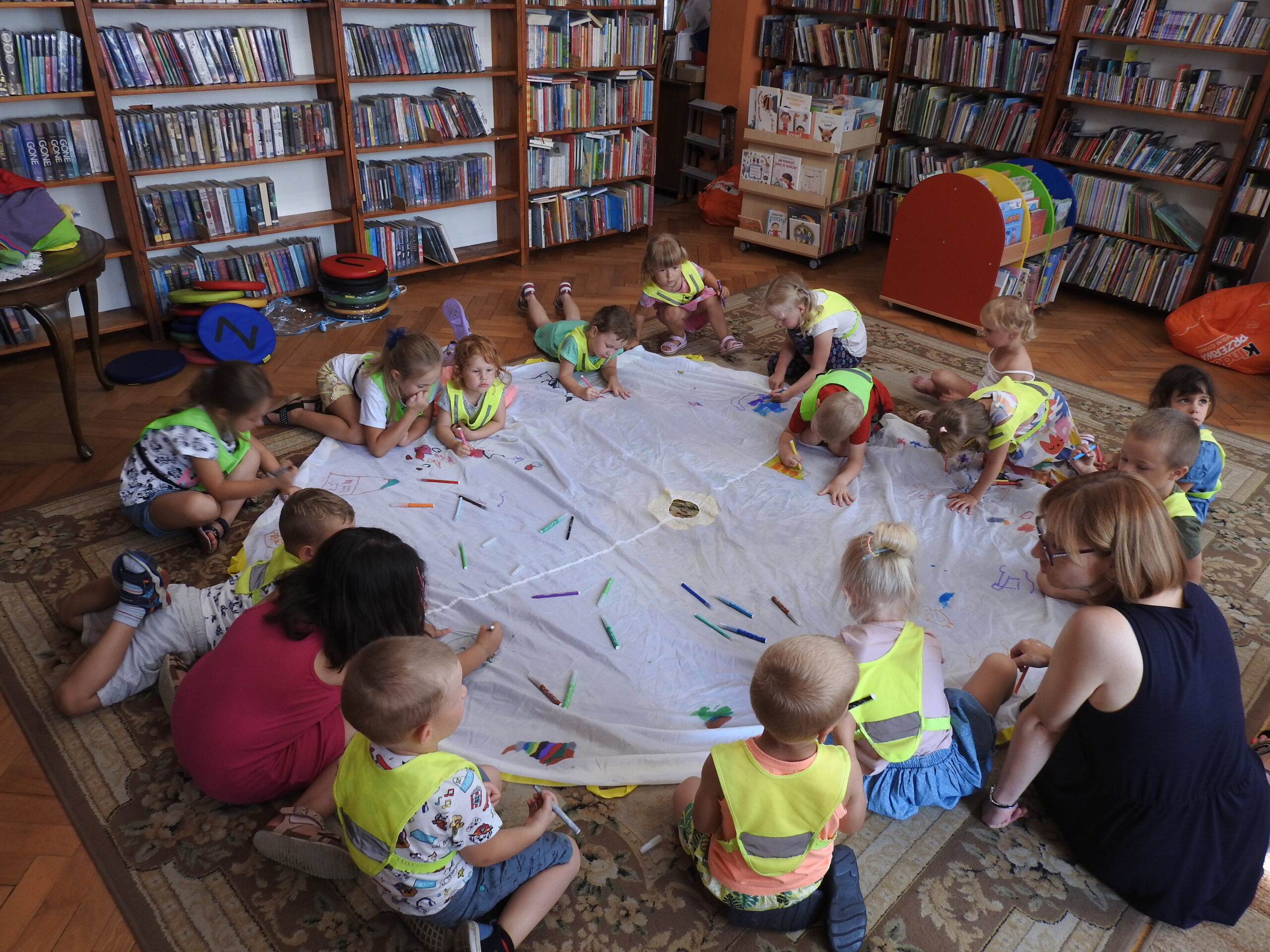 Grupa dzieci w kamizelkach odblaskowych siedzi lub leży na dywanie wokół dużego, białego, okrągłego materiału, na którym leży dużo flamastrów w różnych kolorach. Dzieci malują po materiale. W tle regały z książkami.