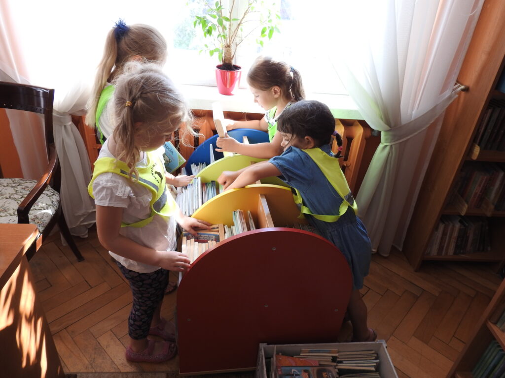 Czworo dzieci stoi wokół małego regału i z zaangażowaniem przegląda znajdujące się na nim książeczki. W tle okno.