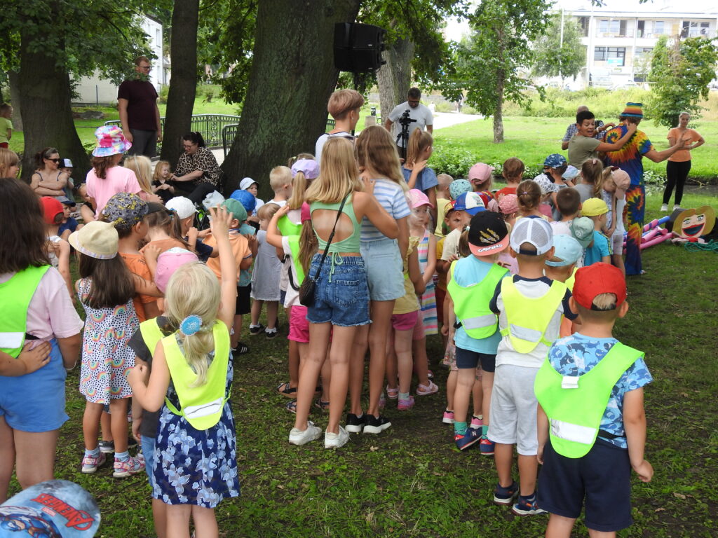 Duża grupa dzieci stoi w kilku szeregach. Dzieci dotykają palcem pleców osoby stojącej przed nimi. Wszystkie dzieci są zwrócone w stronę mężczyzny ubranego w kolorowy strój, który stoi do nich tyłem. Kobieta dotyka jego pleców zwracając głowę w stronę dzieci. W tle drzewa.