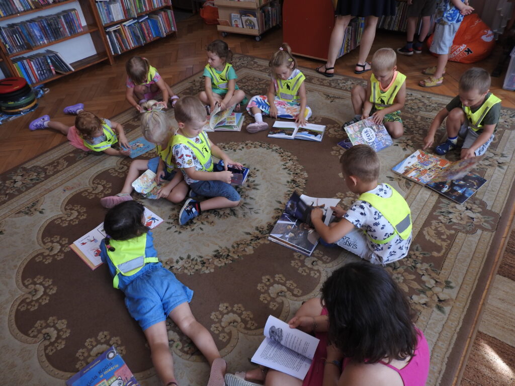 Grupa dzieci w kamizelkach odblaskowych siedzi na dywanie i ogląda książki. W tle regały z książkami.