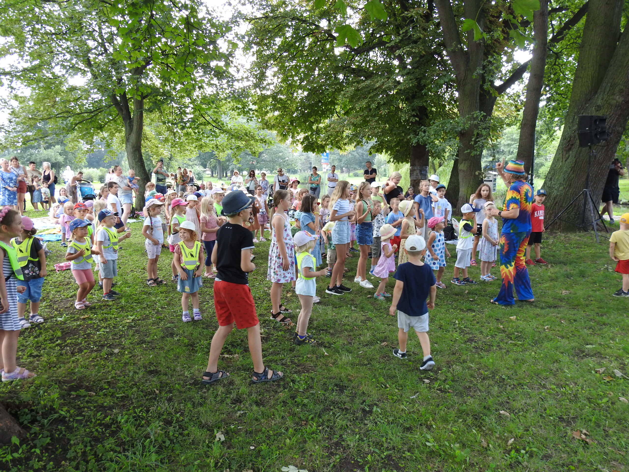 Duża grupa dzieci stoi naprzeciw osoby dorosłej ubranej w kolorowy strój, która trzyma rękę uniesioną w górę. W oddali stoi duża grupa osób dorosłych przyglądających się dzieciom. W tle drzewa.