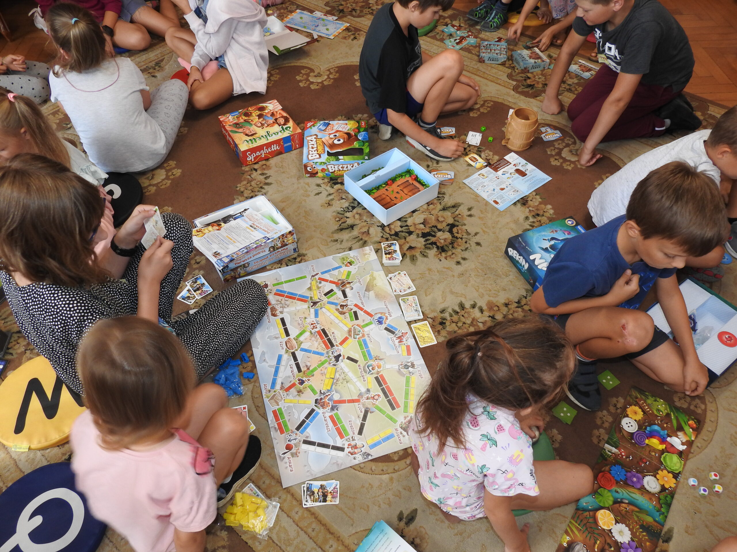 Grupa dzieci siediz na dywanie, na którym są rozłożone otwarte gry planszowe.