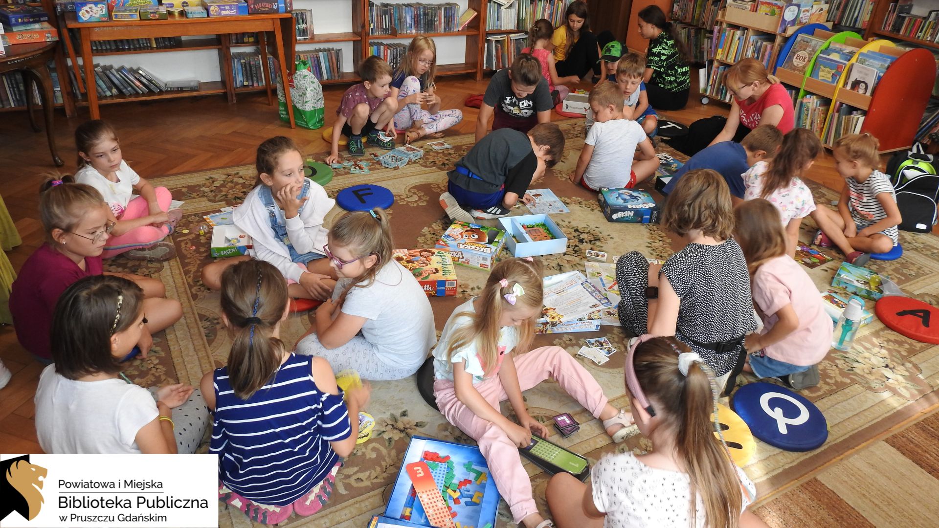 Duża grupa dzieci siedzi na dywanie. Są podzieleni na małe grupki. Przed dziećmi leżą rozłożone różne gry planszowe. W tle regały z książkami.