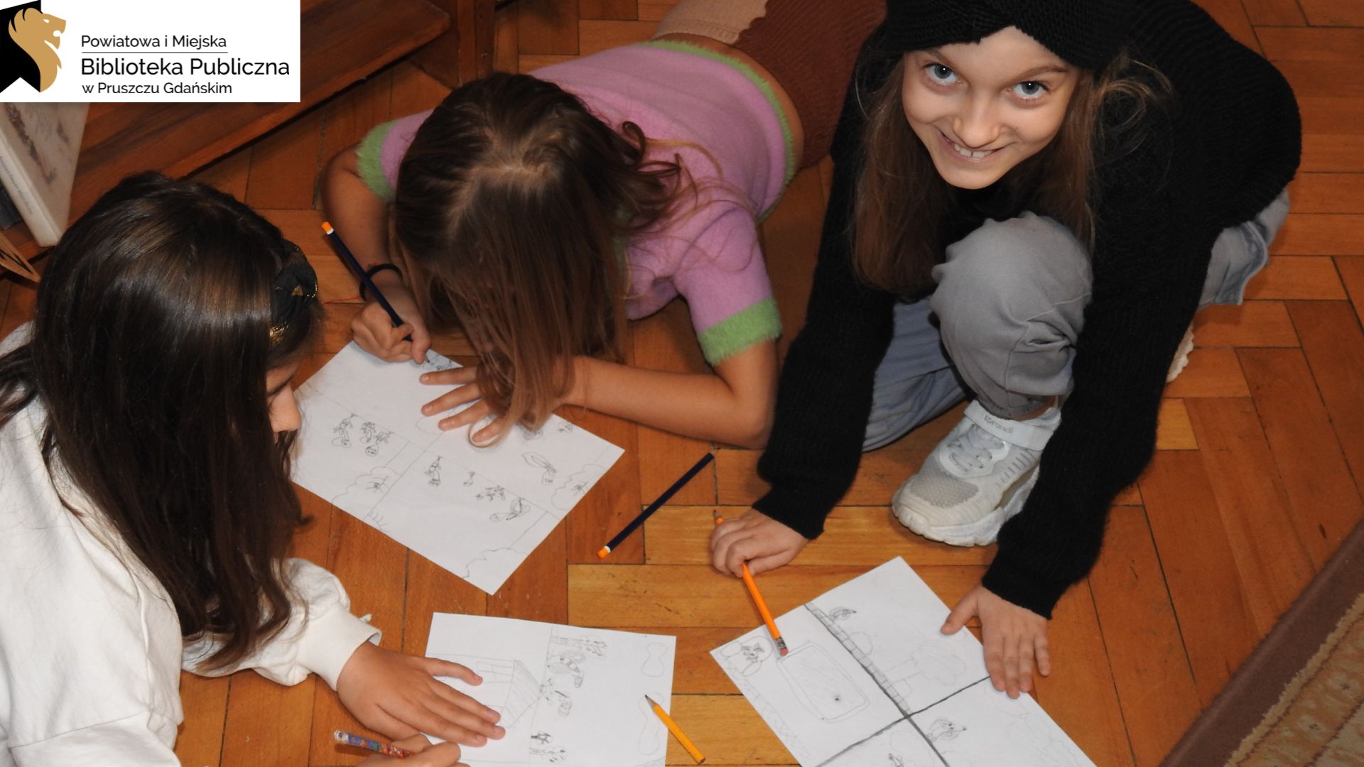 Dwie dziewczynki leżą, a jedna klęczy na podłodze. Przed nimi znajdują się kartki A4 z czarno-białymi szkicami. Dziewczynki trzymają ołówki. Dwie z nich są w trakcie rysowania, a jedna patrzy w górę i uśmiecha się.