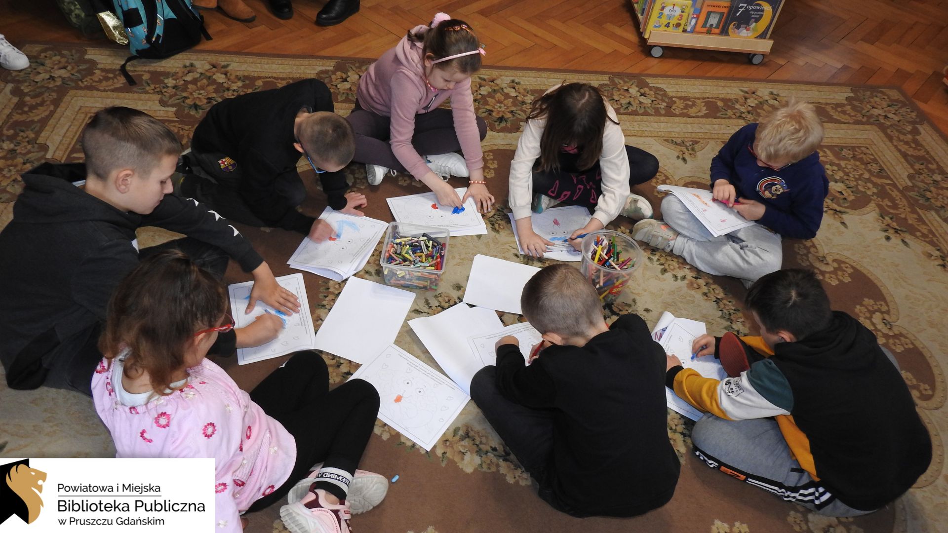 Grupa dzieci siedzi na dywanie w kole. Na środku koła stoją dwa pudełka wypełnione kredkami. Przed dziećmi leżą białe kartki A4 z szablonem do kolorowania. Dzieci wypełniają kolorowankę.