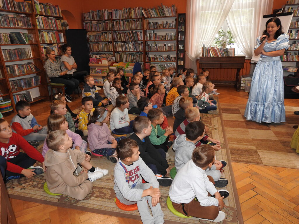 Duża grupa dzieci siedzi na dywanie i jest skierowana w stronę kobiety ubranej w błękitną, bajkową suknię, która trzyma mikrofon przy buzi. W tle regały z książkami.