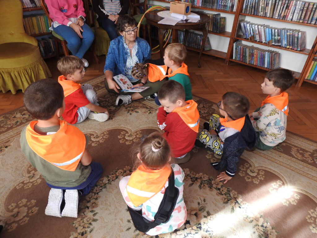 Osoba dorosła oraz ośmioro dzieci siedzą na dywanie. Dzieci są skierowane w stronę osoby dorosłej, która trzyma na kolanach książkę.