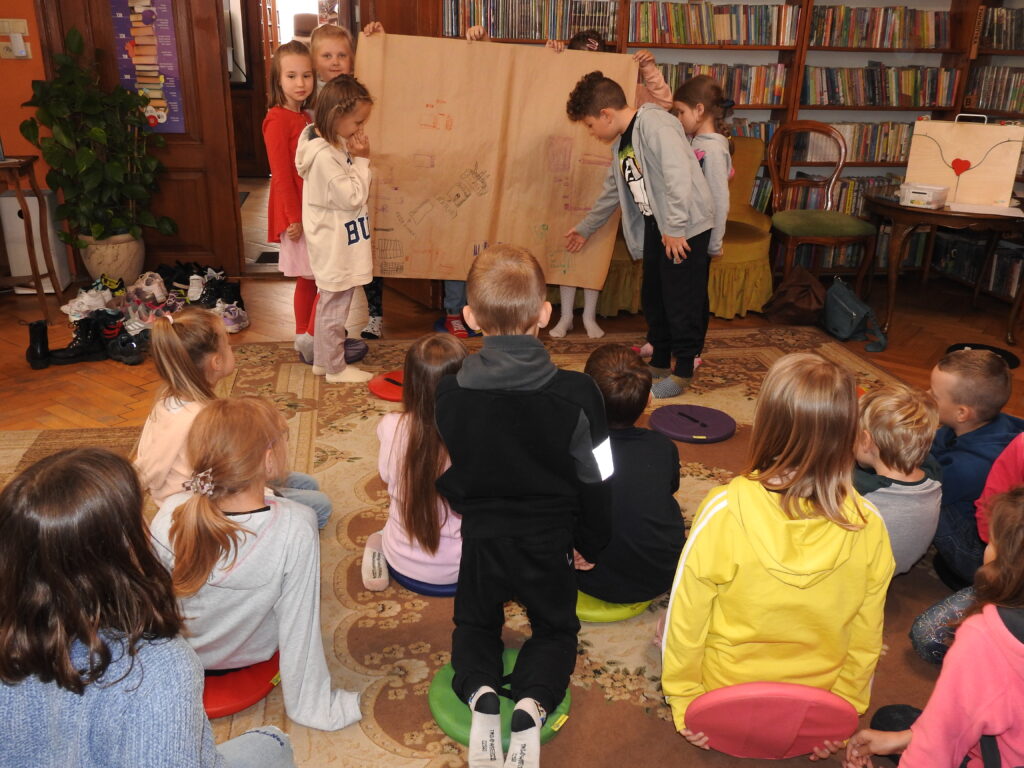 Kilkoro dzieci trzyma duży arkusz szarego papieru z rysunkami dzieci. Jedno dziecko wskazuje ręką na jeden z rysunków. Naprzeciw, na dywanie, siedzi grupa dzieci. Obok stojących dzieci znajduje się stół, na którym jest zamknięta skrzynia teatrzyku kamishibai. W tle regały z książkami.