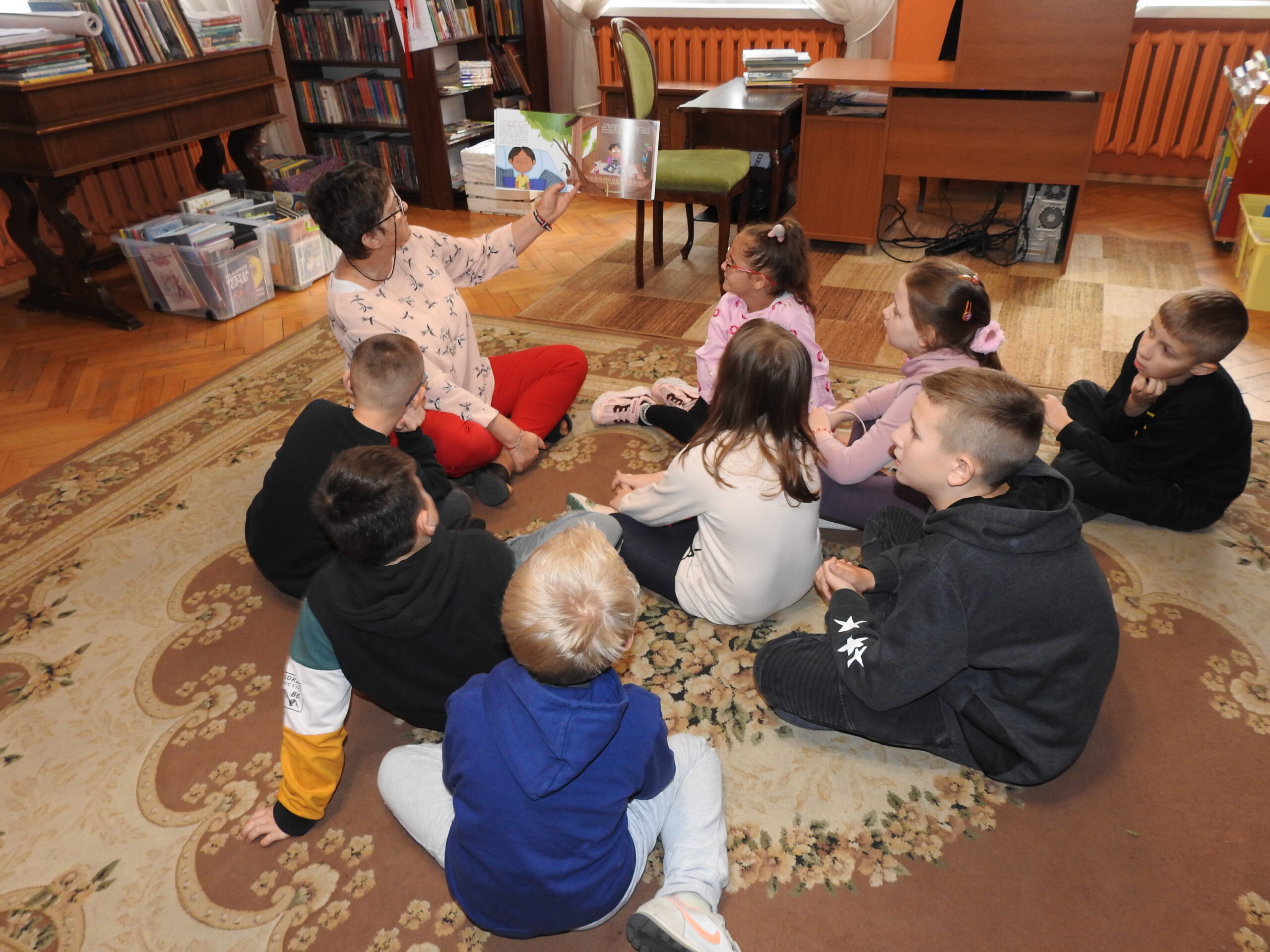 Grupa dzieci siedzi na dywanie. Jest  skierowana w stronę osoby dorosłej, która również siedzi na dywanie. Trzyma otwartą książkę wysoko w lewej ręce, tak by dzieci mogły zobaczyć ilustracje. W tle biurko, sekretarzyk oraz regały z książkami.
