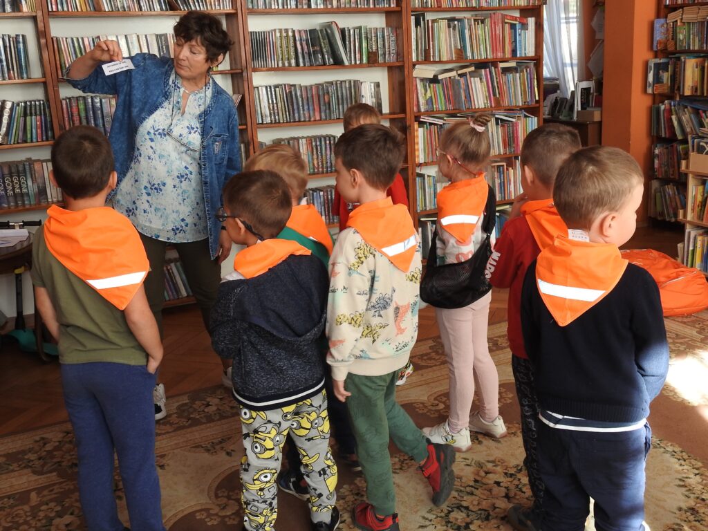 Osoba dorosła stoi na tle regałów z książkami. Trzyma wysoko w ręce małą karteczkę z krótkim, nadrukowanym tekstem. Grupa dzieci stoi naprzeciw niej i patrzy na karteczkę.