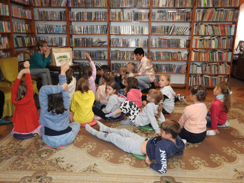Grupa dzieci oraz jedna osoba dorosła siedzą na dywanie lub podłodze. Są skierowani w stronę osoby dorosłej siedzącej na krześle. Obok niej stoi stół, na którym znajduje się zamknięta skrzynia teatrzyku kamishibai. Oparta jest o nią jedna z kart teatrzyku, przedstawiająca budynek ze słomianym dachem. Część dzieci ma uniesione ręce do góry. W tle regały z książkami.