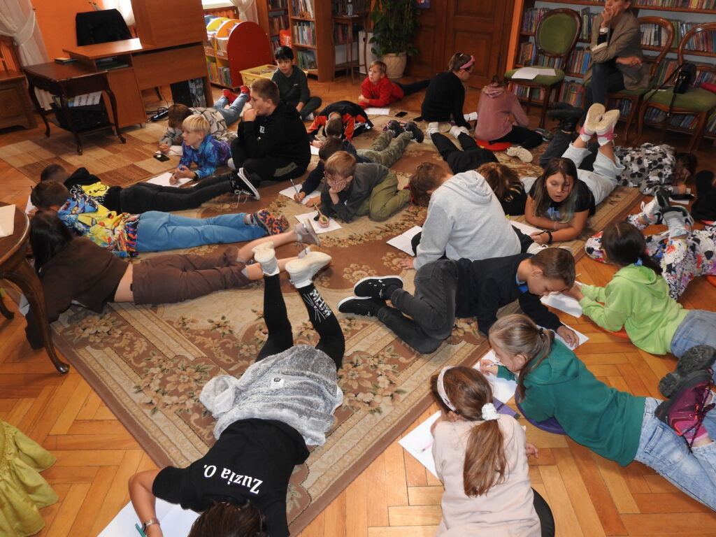 Duża grupa dzieci leży lub siedzi na dywanie i podłodze. Przed nimi leżą białe kartki, na których dzieci rysują ołówkami. W tle biurko oraz regały z książkami.