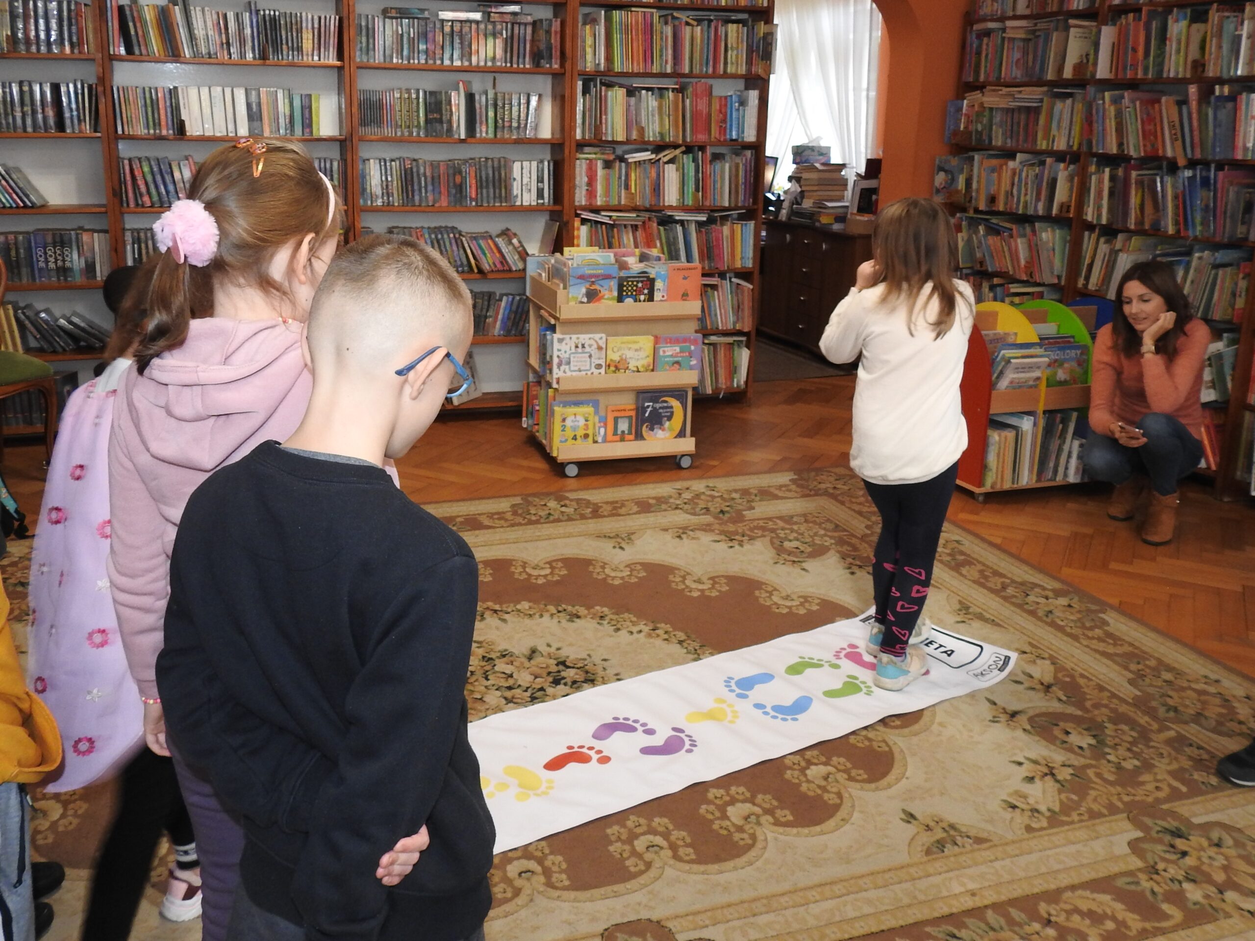 Na dywanie leży podłużna mata z ilustracjami kolorowych stóp. Przed nią stoi kilkoro dzieci. Jedno dziecko znajduje się na macie. Jego stopy dopasowują się do tych nadrukowanych. Naprzeciwko dziecka kuca jedna osoba dorosła. W tle regały z książkami.