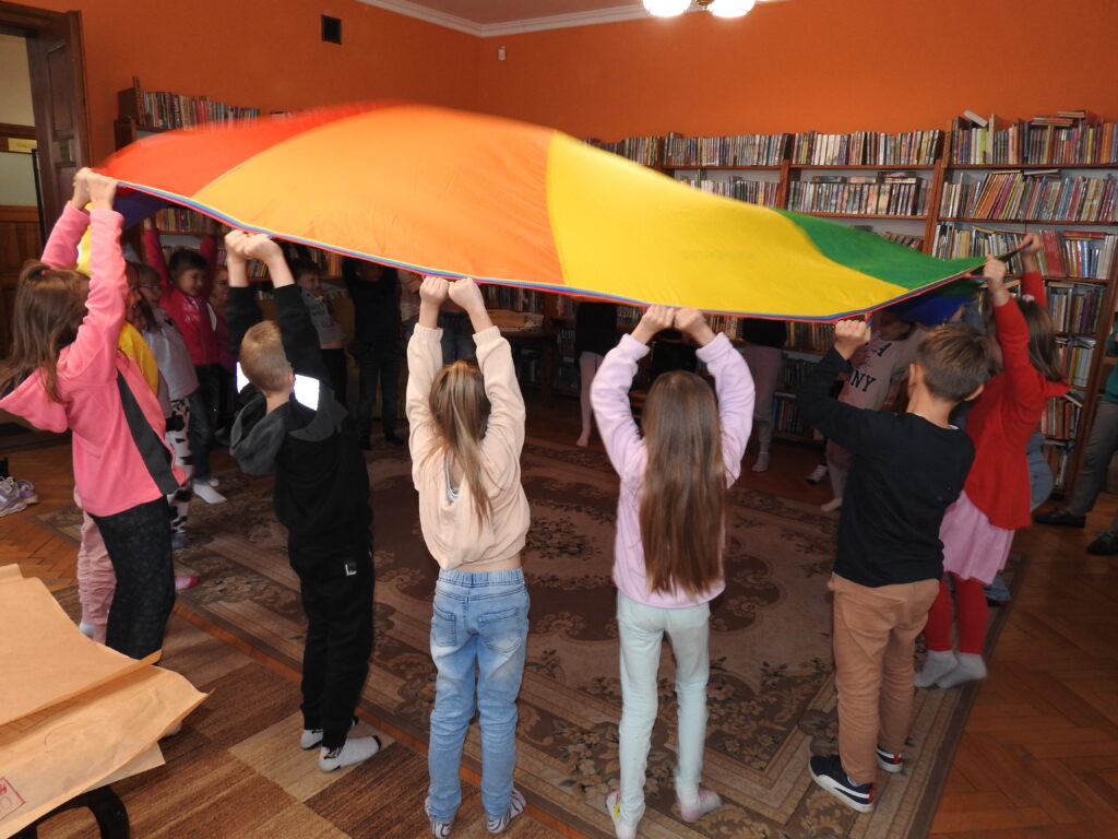 Duża grupa dzieci stoi w kole trzymając nad sobą kolorową chustę animacyjną. W tle regały z książkami.