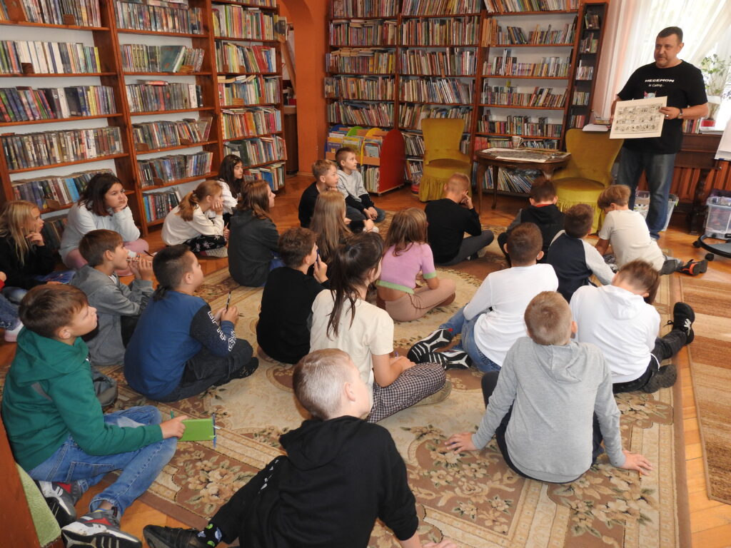 Duża grupa dzieci siedzi na dywanie i patrzy w stronę Bogdana Ruksztełło-Kowalewskiego, który trzyma przed sobą kartę z komiksami. Obok niego stoi stół, na którym znajduje się dużo plansz komiksowych. W tle regały z książkami.