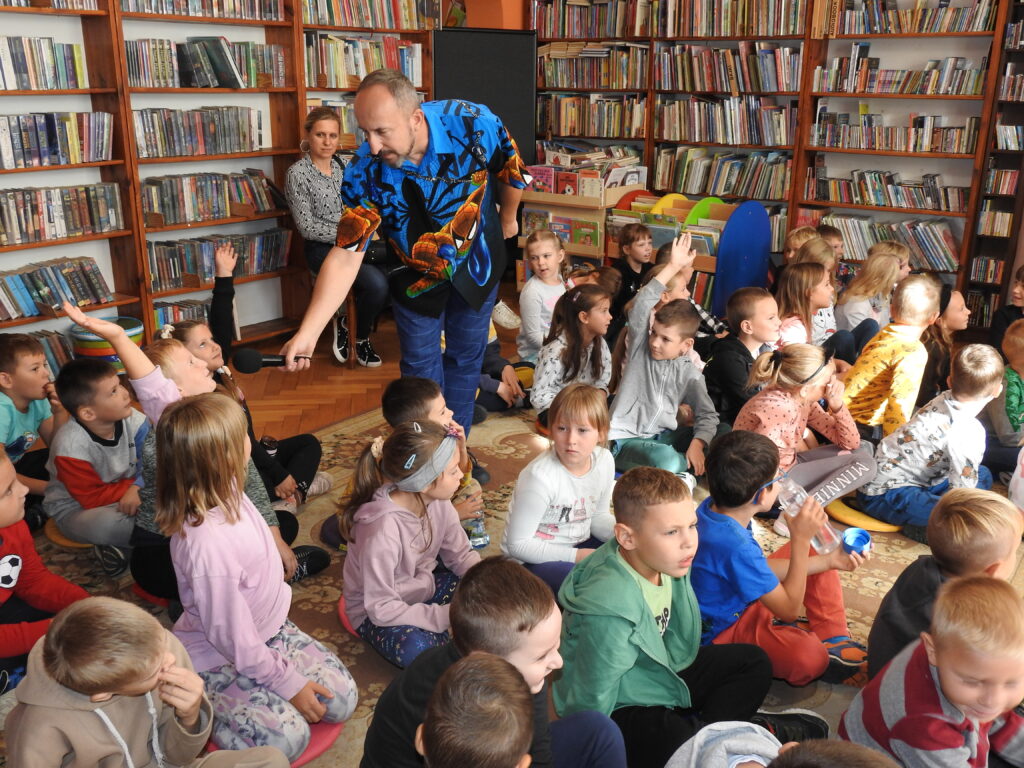 Duża grupa dzieci siedzi na dywanie. Pomiędzy nimi stoi mężczyzna, który pochyla się z mikrofonem w stronę dziecka trzymającego rękę w górze. W tle regały z książkami.