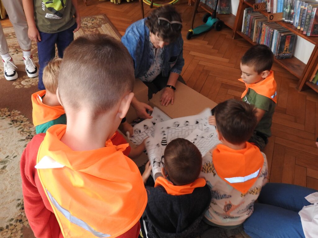 Grupa dzieci oraz jedna osoba dorosła są pochyleni nad kartonowym pudłem, z którego wyjmują kartki z nadrukowanymi labiryntami. 