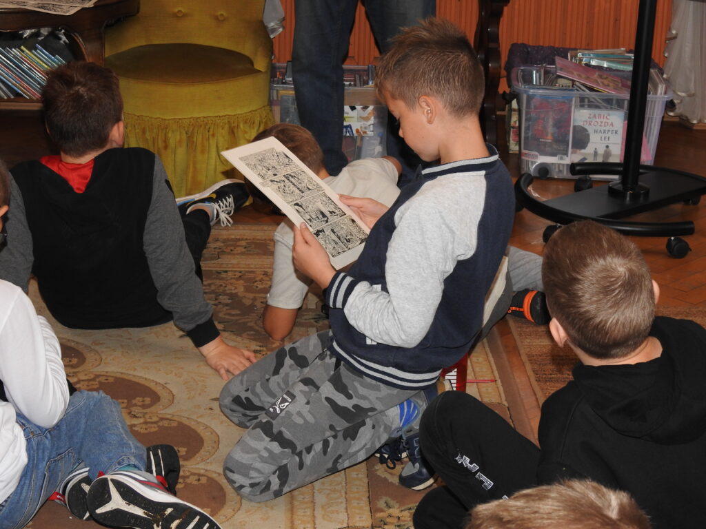 Chłopiec klęczy na dywanie i patrzy na trzymaną przez siebie planszę z komiksami. Obok niego siedzi kilkoro dzieci. 
