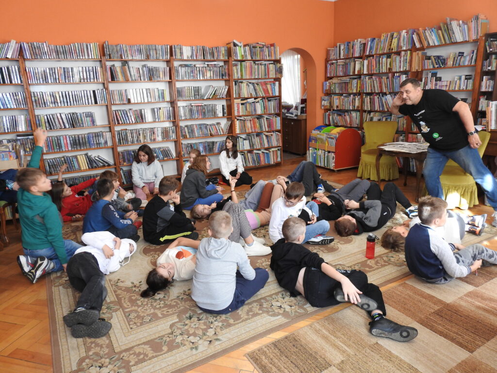 Grupa dzieci siedzi na dywanie i jest skierowana w stronę Bogdana Ruksztełło-Kowalewskiego. Mężczyzna nachyla się w stronę dzieci trzymając się za jedno ucho w geście nasłuchiwania. Kilkoro dzieci ma uniesione ręce w górze. W tle regały z książkami. 