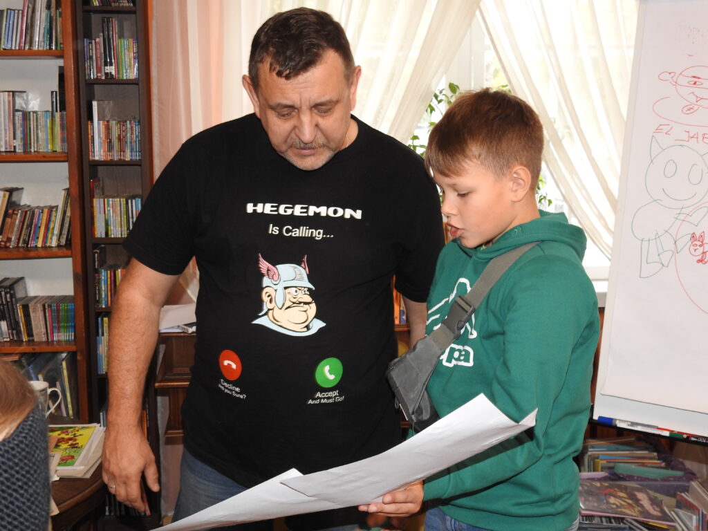Bogdan Ruksztełło-Kowalewski stoi obok chłopca, który trzyma dwie białe kartki A4. Patrzą na rysunki umieszczone na kartkach. 