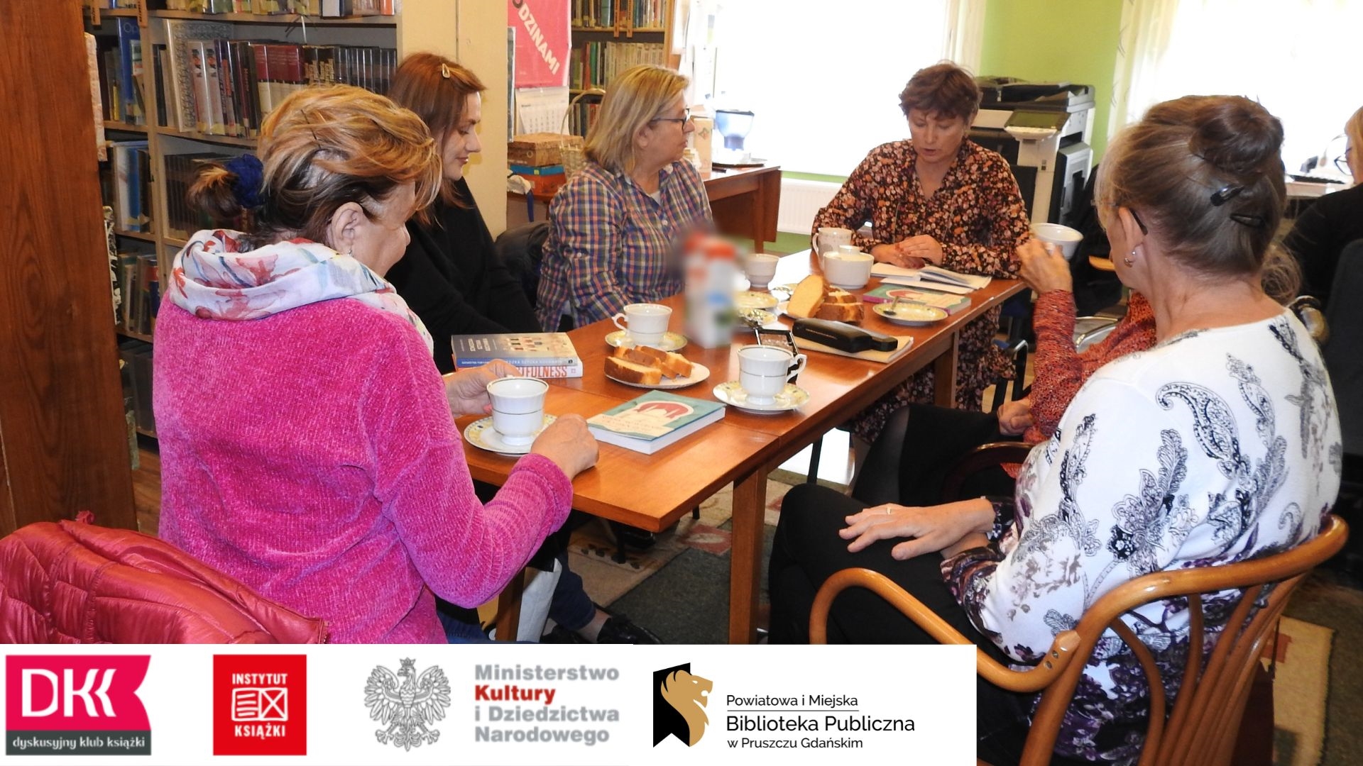 Członkinie Dyskusyjnego Klubu Książki dla dorołsych podczas spotkania we wrześniu (5 osób) oraz moderatorka Anna Krawycińska. Na stole leżą książki, kartki z notatkami oraz stoją filiżanki z kawą, ciastka i ciasto.