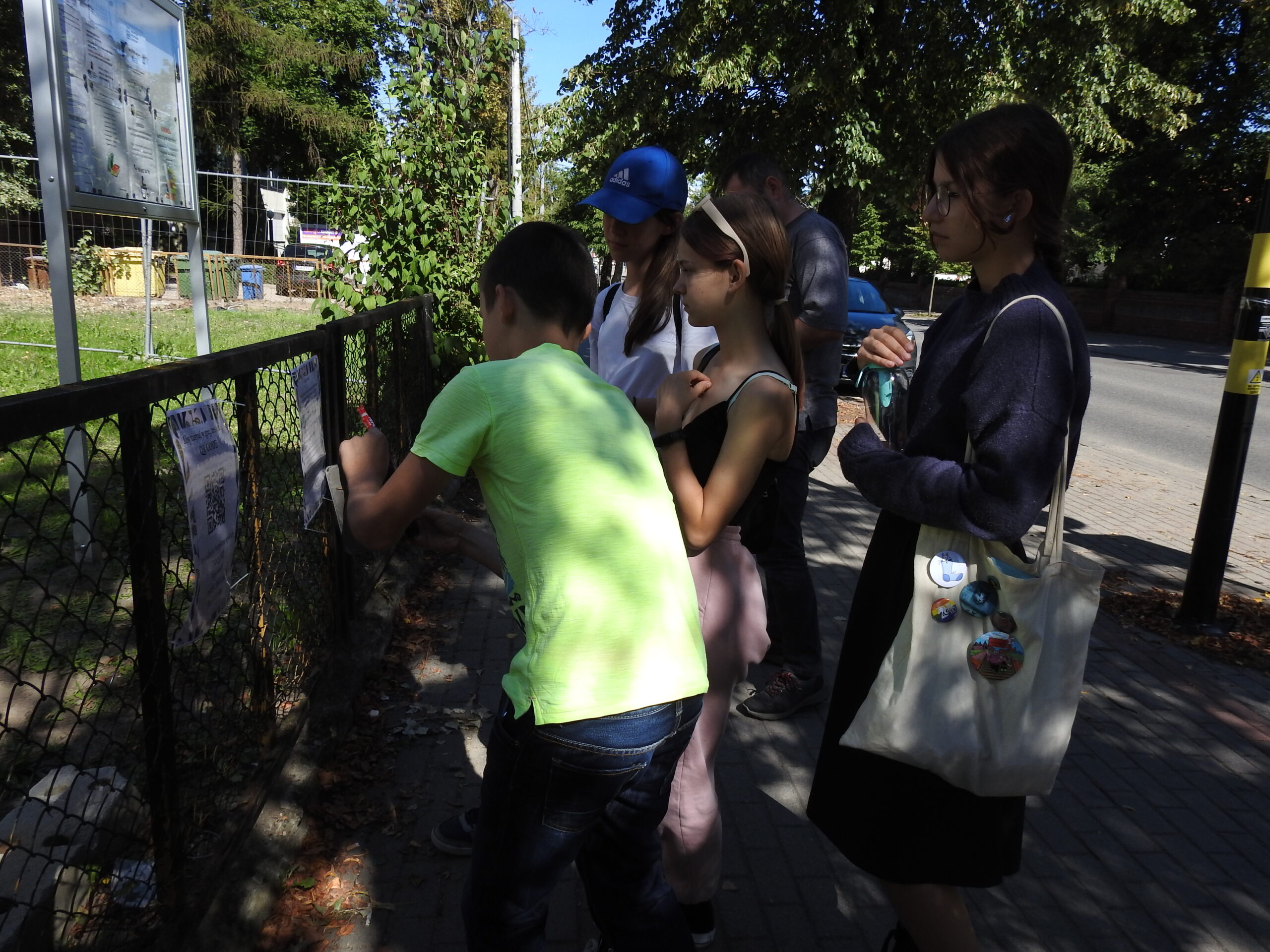Grupa nastoletnich dzieci stoi przed  ogordzeniem wiodącym do biblioteki, na kórym wiszą zasady gry oraz kod QR. Jedna z osób robi zdjęcie telefonem komórkowym.