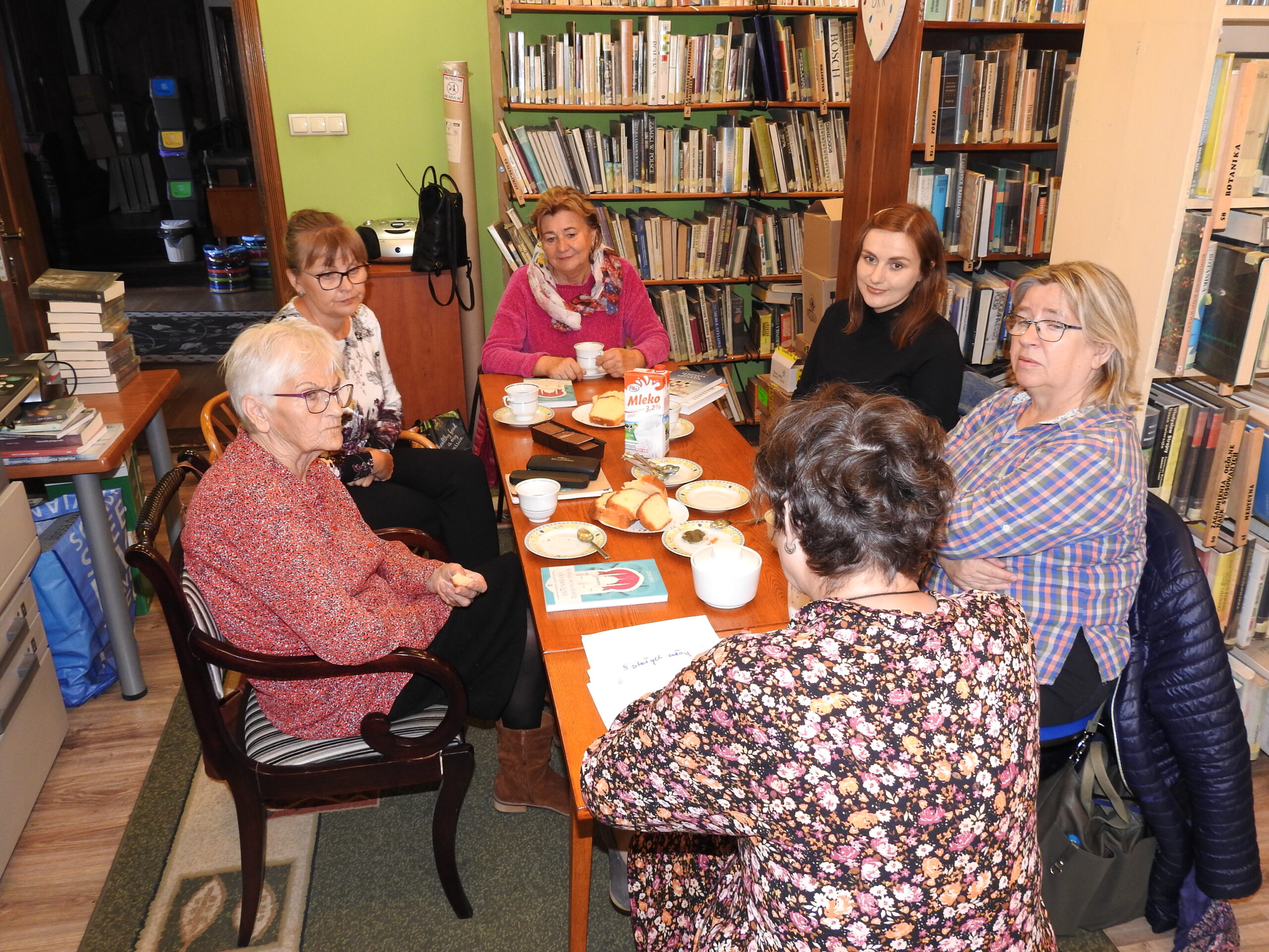 Moderatorka oraz członkinie Dyskusyjnego Klubu Książki(6 osób) siedzą przy stole i rozmawiają o książce. Na stole leżą ksiązki, stoja filiżanki z napojem, ciastka.