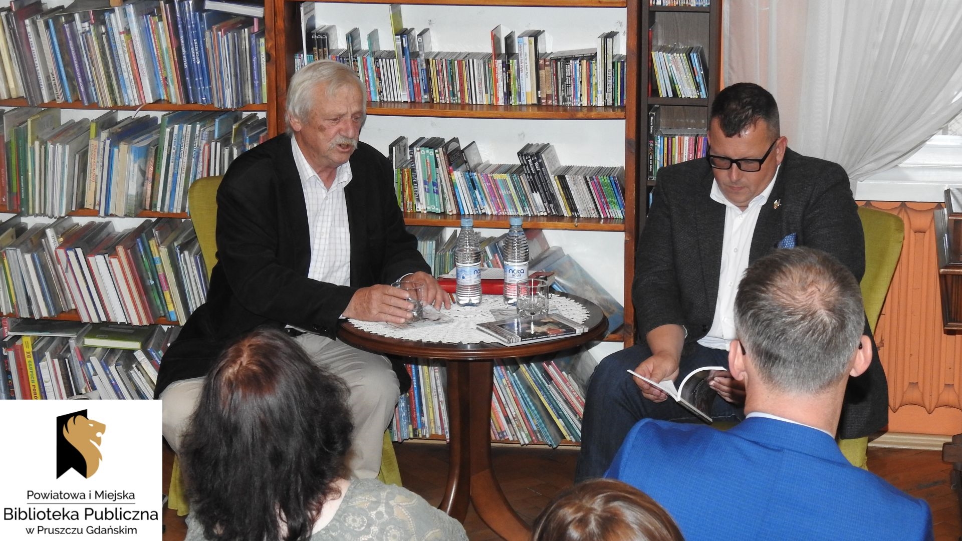 Wawrzyniec Rozenberg oraz Bartosz Gondek siedzą przy stoliku. Za mężczyznami regały z książkami.