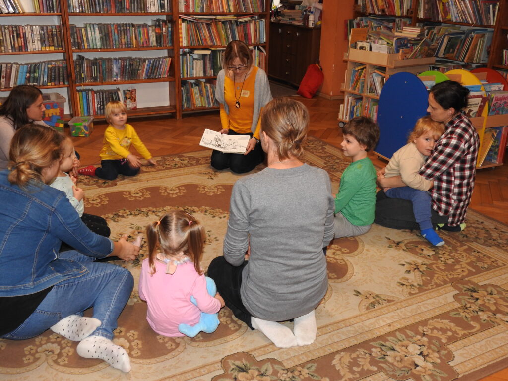 Pięcioro małych dzieci oraz pięcioro dorosłych siedzi lub klęczy w kole na dywanie. Jedna z osób, bibliotekarka, trzyma na kolanach otwartą książkę skierowaną obrazkiem do dzieci. W tle regały z książkami.