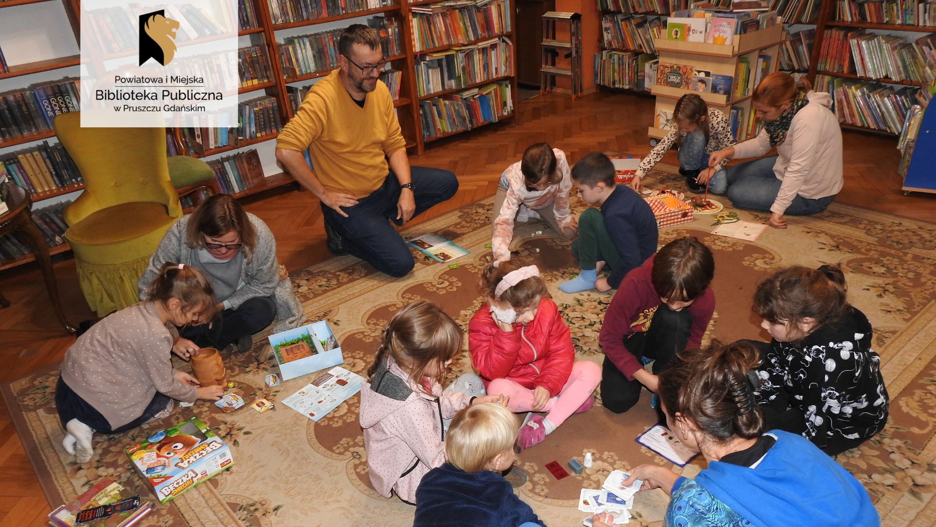 Dziewięcioro dzieci oraz cztery osoby dorosłe siedzą lub kucają na dywanie. Tworzą cztery grupy. Przed każdą z grup leży rozłożona gra planszowa. W tle regały z książkami.
