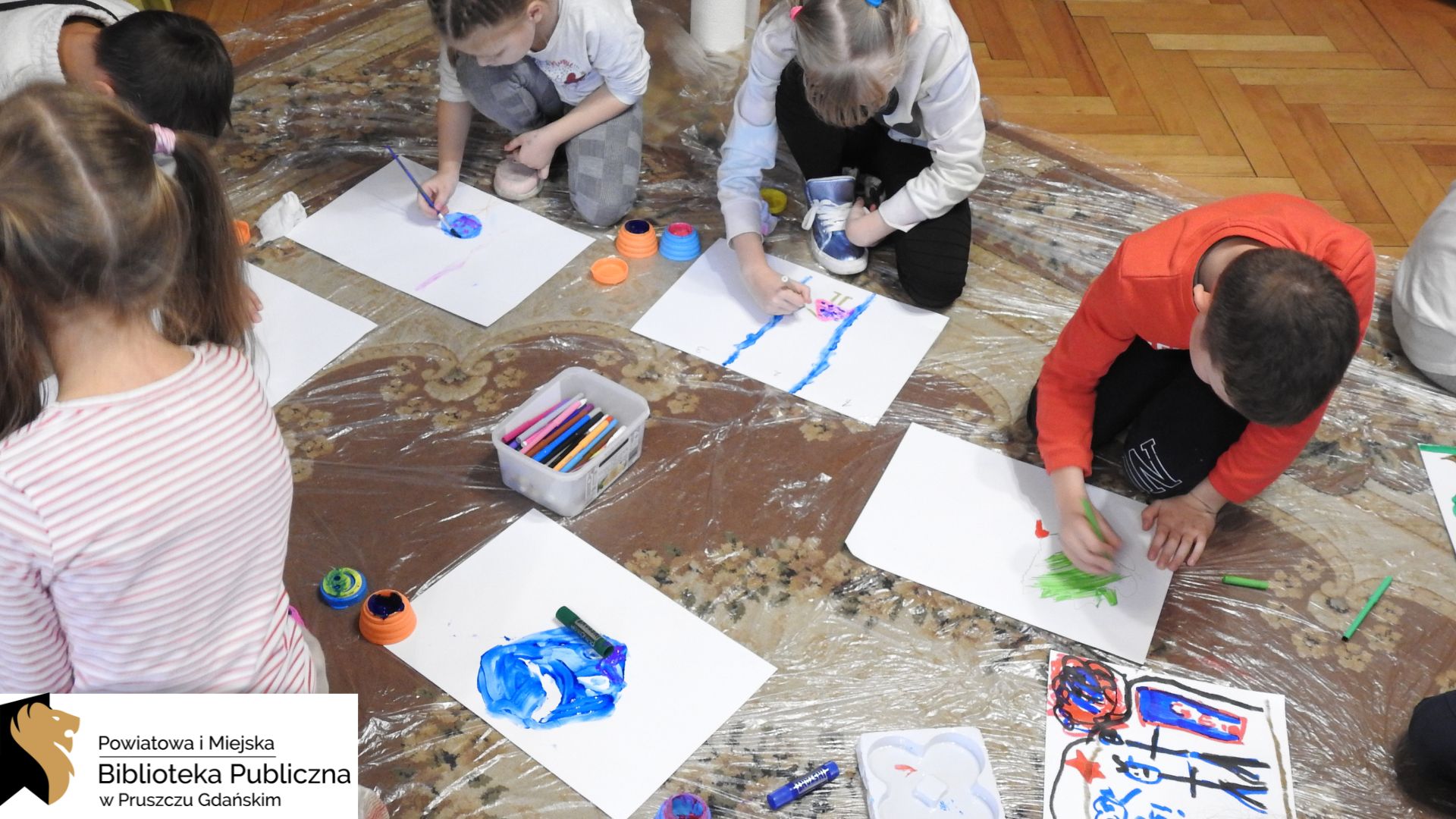 Grupa dzieci klęczy na dywanie wyłożonym przezroczystą folią. Dzieci trzymają pędzelki i malują farbami na leżących przed nimi białych kartkach formatu A3. Pomiędzy nimi znajdują się kolorowe pudełeczka z farbami oraz pudełko z flamastrami. Na kartkach utworzone są już fragmenty pracy plastycznej.