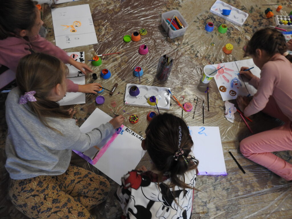 Czworo dzieci siedzi lub klęczy na dywanie pokrytym przezroczystą folią. Przed dziećmi leżą białe kartki formatu A3 oraz wiele kolorowych pudełeczek z farbami. Dzieci malują pędzelkami lub dotykają farb. Na kartkach utworzone są już fragmenty pracy plastycznej. 