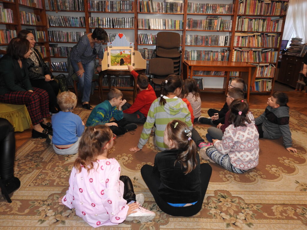 Grupa dzieci siedzi na dywanie. Jest skierowana w stronę otwartego teatrzyku kamishibai. Jedno z dzieci wskazuje palcem na kartę teatrzyku, na której nadrukowana jest gąsienica obok przewróconego pnia. Jedna osoba dorosła pochyla się nad teatrzykiem, a dwie siedzą obok na krzesłach.