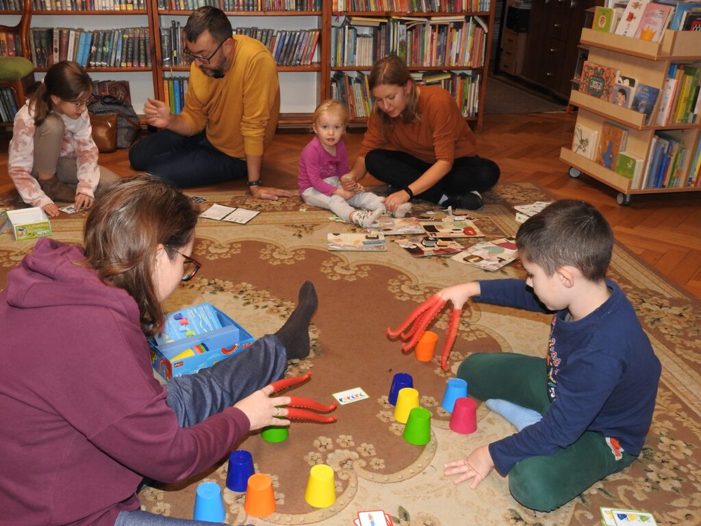 Trzy osoby dorosłe oraz troje dzieci siedzą na dywanie. Dwie osoby mają założone na dłonie czerwone, gumowe macki ośmiornicy i za ich pomocą chwytają kolorowe kubeczki. Pozostałe osoby mają rozłożone przed sobą kolorowe plansze lub karty.