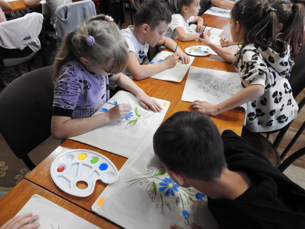 Grupa dzieci siedzi przy stole, na którym znajdują się płócienne torby oraz palety z farbami. Dzieci kolorują kwiatowy kaszubski wzór na torbach.