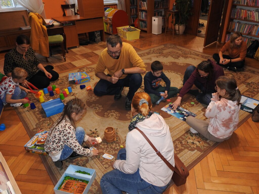 Pięć osób dorosłych oraz czworo dzieci siedzi na dywanie. Tworzą cztery grupki. Przed każdą z nich leżą rozłożone gry.
