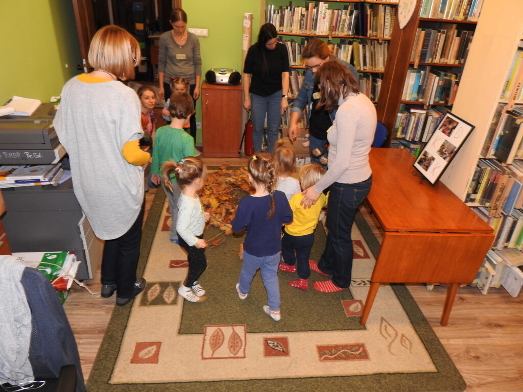 Sześcioro dzieci oraz sześcioro dorosłych stoi wokół dużej ilości liści i patyków rozłożonych na dywanie. W tle regały z książkami oraz sprzęt biurowy.