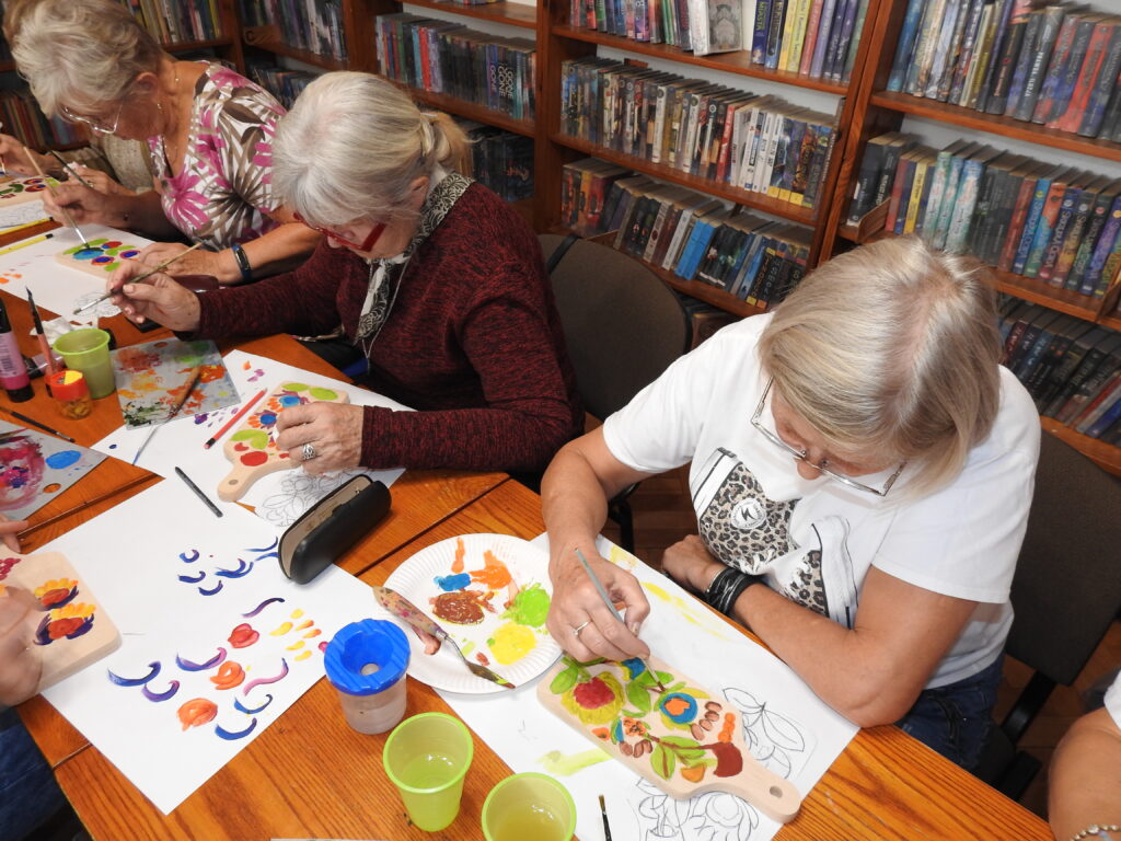 Trzy kobiety siedzą przy stole, na którym leżą drewniane deski, białe kartki formatu A3 częściowo pomalowane farbami, pędzle, pojemniki z wodą, palety do mieszania farb. Kobiety malują pędzelkami na deskach kwiatowe wzory.