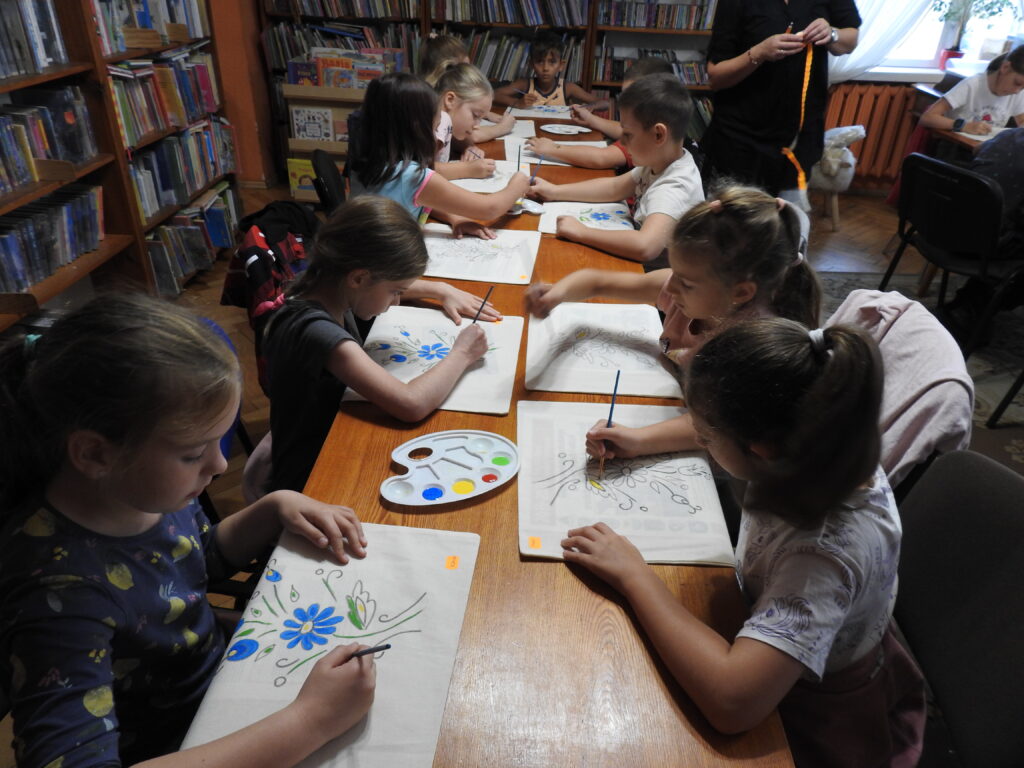 Grupa dzieci siedzi przy stole, na którym znajdują się płócienne torby oraz palety z farbami. Dzieci kolorują kwiatowy kaszubski wzór na torbach.